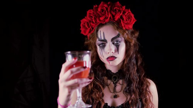 Easy Halloween Makeup. The girl with the picture on her face. The devil's bride with a wreath of red flowers on her head. The woman is wearing a black corset dress and black stockings. Woman drinks