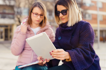 Two women female friends using digital tablet notebook in the city looking to the pictures or map application project in winter or autumn day talking discussing work