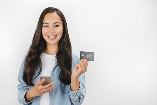 Portrait of smiling asian woman holding credit card and mobile phone while looking at camera isolated over white background