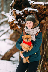 Young positive girl in winter clothes holding in hands her adorable pomeranian puppy in snowy forest
