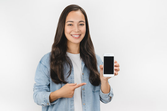 Happy Young Asian Woman Holding Blank Screen Mobile Phone And Pointing Finger Over White Background