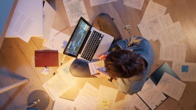 Flat lay of young woman sitting on floor surrounded with papers with laptop on her knees, working on it and making notes in notebook in her hands