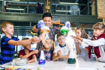 Asian young teacher with group of six cheerful caucasian 8-10 years pupils wearing protective glasses during chemical experiment with colored liquids in modern school.