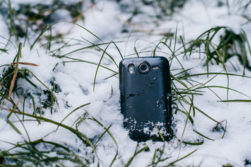 Smaprtphone lying in winetr snowy field at nature with screen covered with snowflakes.