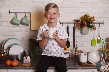 A handsome blond boy is sitting on the kitchen table with a big white cup in his hand. Funny photo