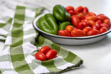 Freshly picked red grape tomatotes and green bell peppers in a white bowl on white marble countertop with green and white checked kitchen towel.