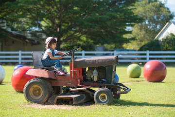 Cute asian little girl playing by driving a old tractor on the green lawn in the farm with the background of green trees, concept of learning by playing for kid development.