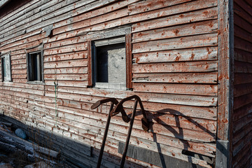 A red shed has worn and red fading paint. It has three boarded up windows and two rusty anchors leaning against the building. The sun is shining on the textured building exposing rusty nail heads.
