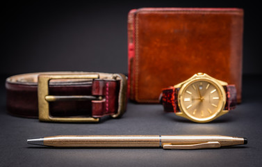 Gold pen with brown leather wallet, red belt and watch isolated on black gradient background. Gift concept for men. Luxury accessories.
