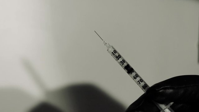 Small clear syringe with liquid inside being squirted by a hand with a black glove on.