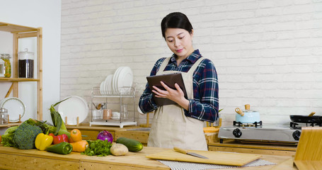 young asian japanese girl in apron using digital tablet device while cooking in kitchen at home. happy smiling woman preparing healthy meal and searching vegan recipes via portable computer indoors.