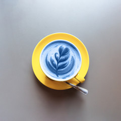 Flat lay of yellow hot trendy latte in blue color with art flower petals on the foam on grey table background. Color of the Year 2020.