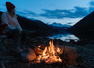 Young woman sitting near campfire next to lake in the evening after sunset with mountains in background in Canada - 307542493