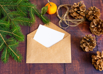 Obraz na płótnie Canvas Blank white paper card in a brown envelope from handmade craft paper on an old wooden table in a Christmas style. Fir branches, mandarin and cones are on the table. Christmas and New Year..