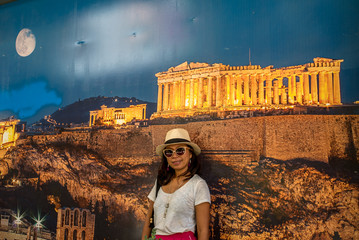 Asian woman in white t-shirt in Athens in white hat, standing in front of large poster of Acropolis...