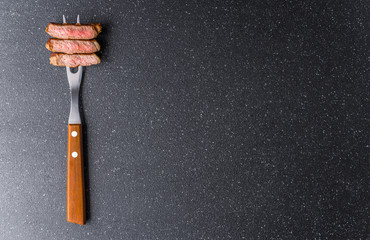 New york strip loin beef steak on a meat fork against black stone background