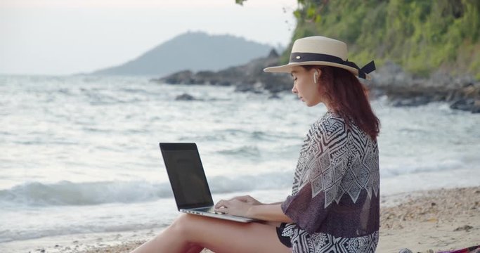 cute dark-haired girl in white hat and tunic rests on sand beach, typing emails