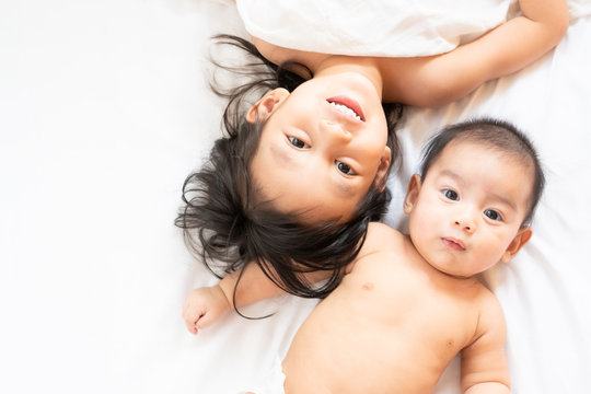 Asian big sister and little baby brother lying on white bed and they are smiling with happy moment in the morning time, concept of love and relation in family, top view photograph.