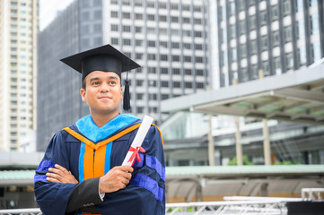 Happy graduate. Happy Asain man in graduation gowns holding diploma in hand on urban city...
