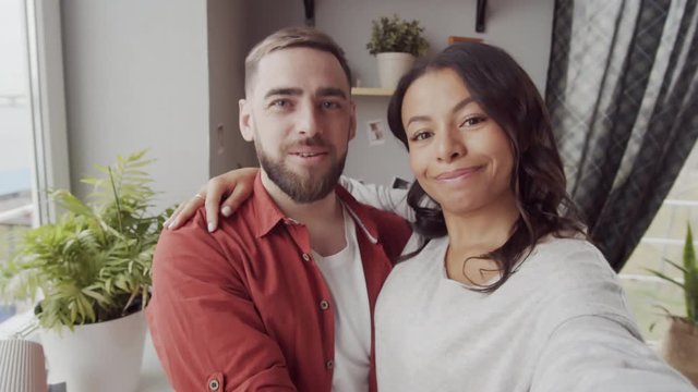Chest-up handheld shot of happy Caucasian husband and mixed race wife with outstretched hand standing by window at home, hugging, waving hello and video chatting with someone