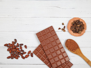Bars of milk chocolate, carob, cocoa powder and cocoa beans on light wooden background
