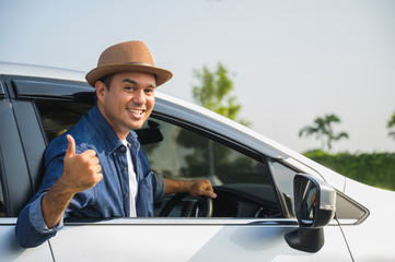 A traveler wearing a hat is driving a car and then pulling himself out of the car window. And thumbs up This picture is about a safe journey by car.