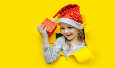 A funny little smiling girl with eyes closed in a red Santa hat looks out of a hole in yellow paper and holds a box with a gift in her hand. Christmas and New Year concept. Background, copy space.