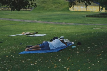              Leisure park in Perth, Western Australia. A woman is lying on green grass. Summer relax. Picnic. Horizontal photograph. Straw hat, white t shirt and blue carpet.          