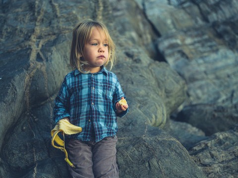 Little toddler eating banana by rocks
