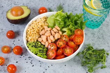 A plate of fresh salad with white beans, bulgur, cherry tomatoes and avocado, decorated with black sesame seeds with products around the plate. Horizontal photo with copy space, top view