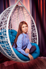 Beautiful brunette girl in the blue shirt on a white wicker hanging chair with pillow in the room with curtains. Model during photoshoot in studio