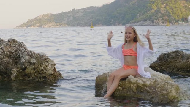 Cute girl teenager shaking throw long hair on sea stones. Beautiful girl in swimsuit and tunic shaking long hair for photo in sea water on stony beach