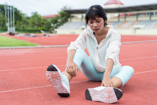 Tired sporty Asian girl sitting down on track after workout in stadium area. Young female hands touch suffering podalgia,feeling pain, ankle or foot broken or twisted. Sport accident and foot sprain