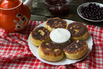 syrniki with a golden crust on a white plate, sour cream and berries, currants. Wooden table and checkered tablecloth. 