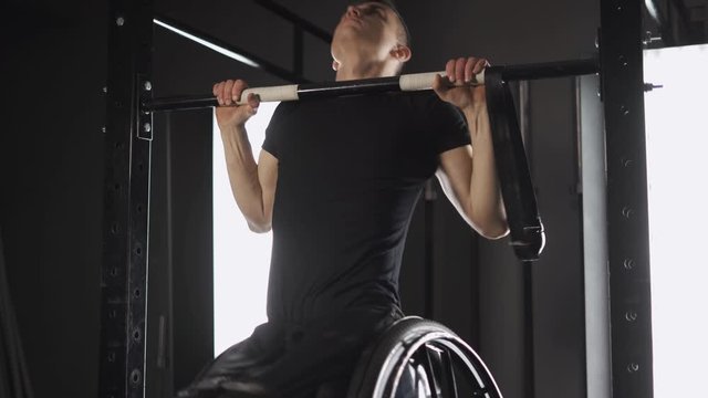 Workout, young disabled athlete in wheelchair performs pull-ups on the horizontal bar, gym training and physical activity, people with disabilities.