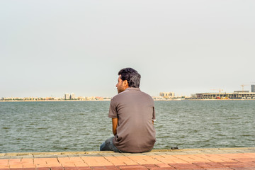 a lonely man is sitting on the rocks after divorce at seaside wearing grey t shirt and blue jeans in the bright cloudy day