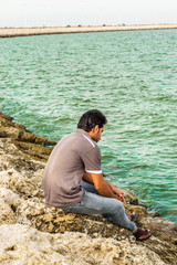 a lonely man is sitting on the rocks after divorce at seaside wearing grey t shirt and blue jeans in the bright cloudy day