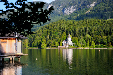 A large white house in the form of a castle on the shore of the lake. Green mountains in the background.