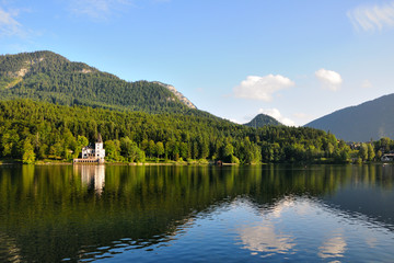 Fototapeta premium A large white house in the form of a castle on the shore of the lake. Green mountains in the background. Blue sky, summer day.