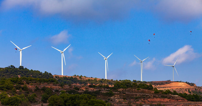 Rows of modern windmills in an energy wind farm on top of a hill against a blue sky and four paragliders in the air