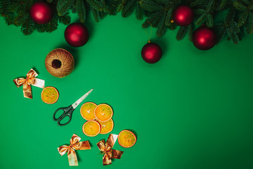 Christmas hand made how to make Christmas wreath. Scissors, rope, red christmas balls, golden bows with fir branches and dried oranges on the green background.