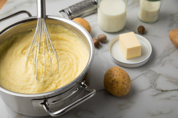 Kochtopf mit Kartoffelpüree mit Zutaten aus Kartoffeln, Milch, Salz, Butter, Muskatnuss dazu Muskatreibe und Schneebesen auf hell Marmor Hintergrund