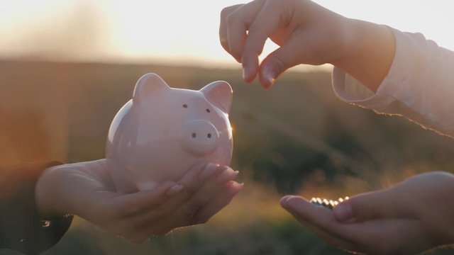 Hands mother and daughter child putting coins into piggy bank. Woman holds piggy bank while girl puts coins at sunset time, lifestyle. Concept kid saving money for future.
