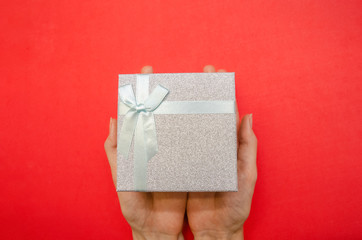 Top view of women's hands holding a shiny gray gift box with a white bow on a red background, copy space