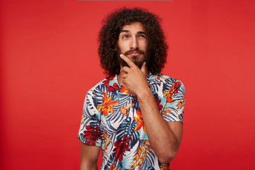 Pensive handsome young brunette curly male with beard holding his chin with raised hand and looking thoughtfully aside, squinting and wrinkling his forehead while standing over red background