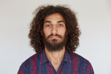 Fototapeta premium Portrait of young handsome brown-eyed bearded male with dark curly hair looking at camera with calm face, keeping lips folded while standing against white background