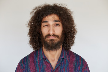 Portrait of pretty young dark haired curly bearded man with folded lips keeping eye closed while looking to camera, wearing casual clothes while standing over white background