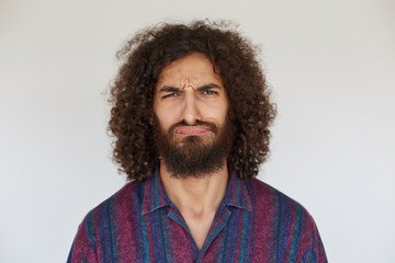 Displeased young pretty bearded male with curly dark hair looking to camera with pout, frowning eyebrows with folded lips against white background, wearing striped casual shirt