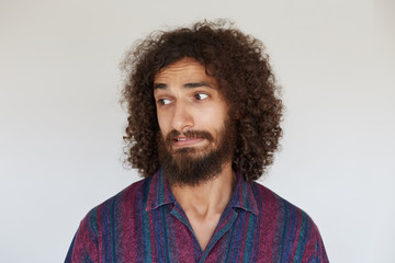 Studio shot of handsome young brunette curly man with beard looking aside with confused face and twisting his mouth, dressed in striped multi-colored shirt against white background