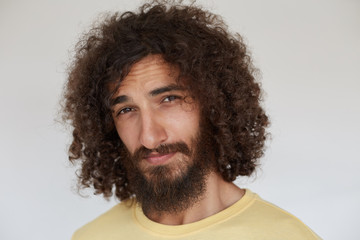 Studio photo of flirty young brunette curly man with beard looking playfully at camera with folded...